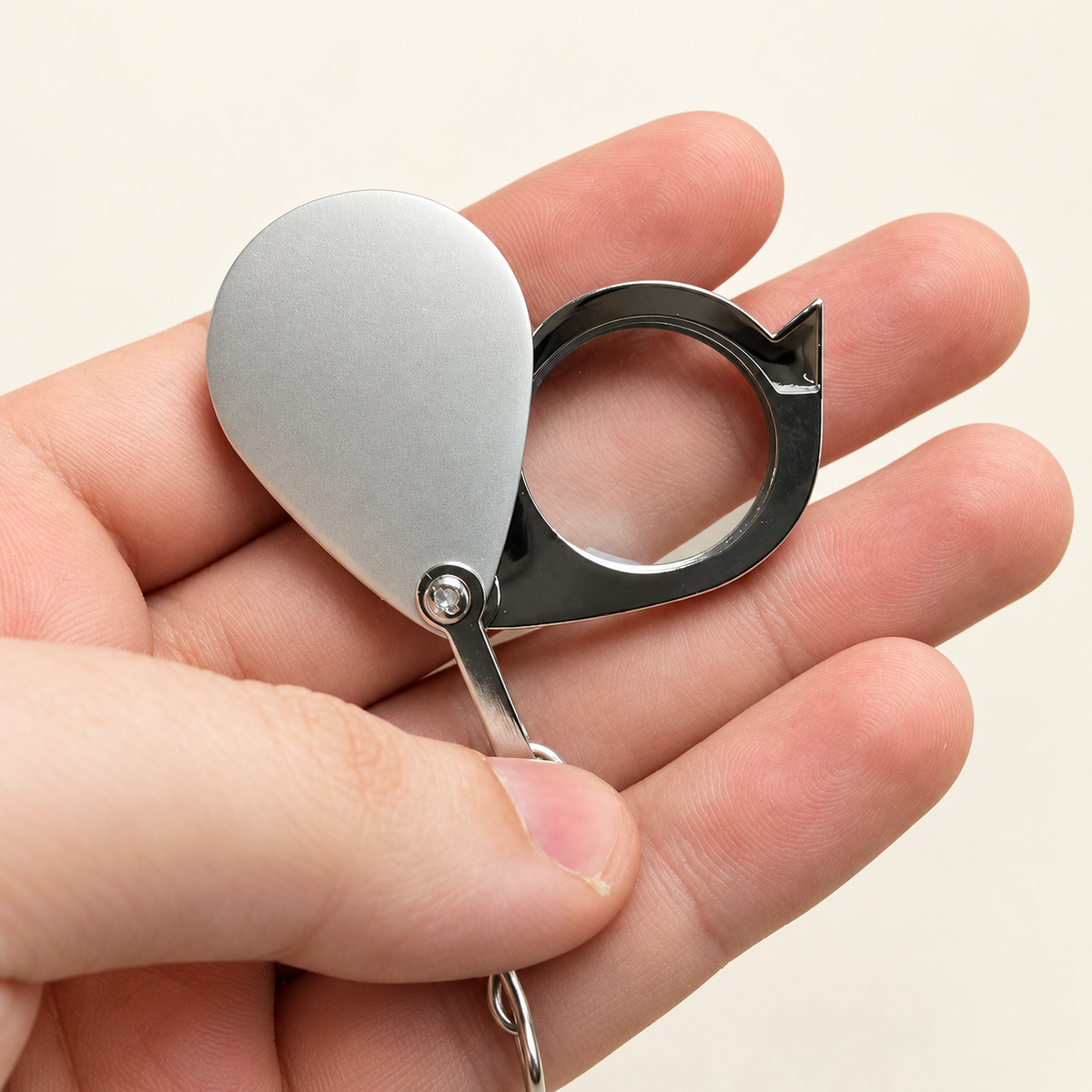 Open silver-tone folding jeweler’s loupe held in a bare hand for scale