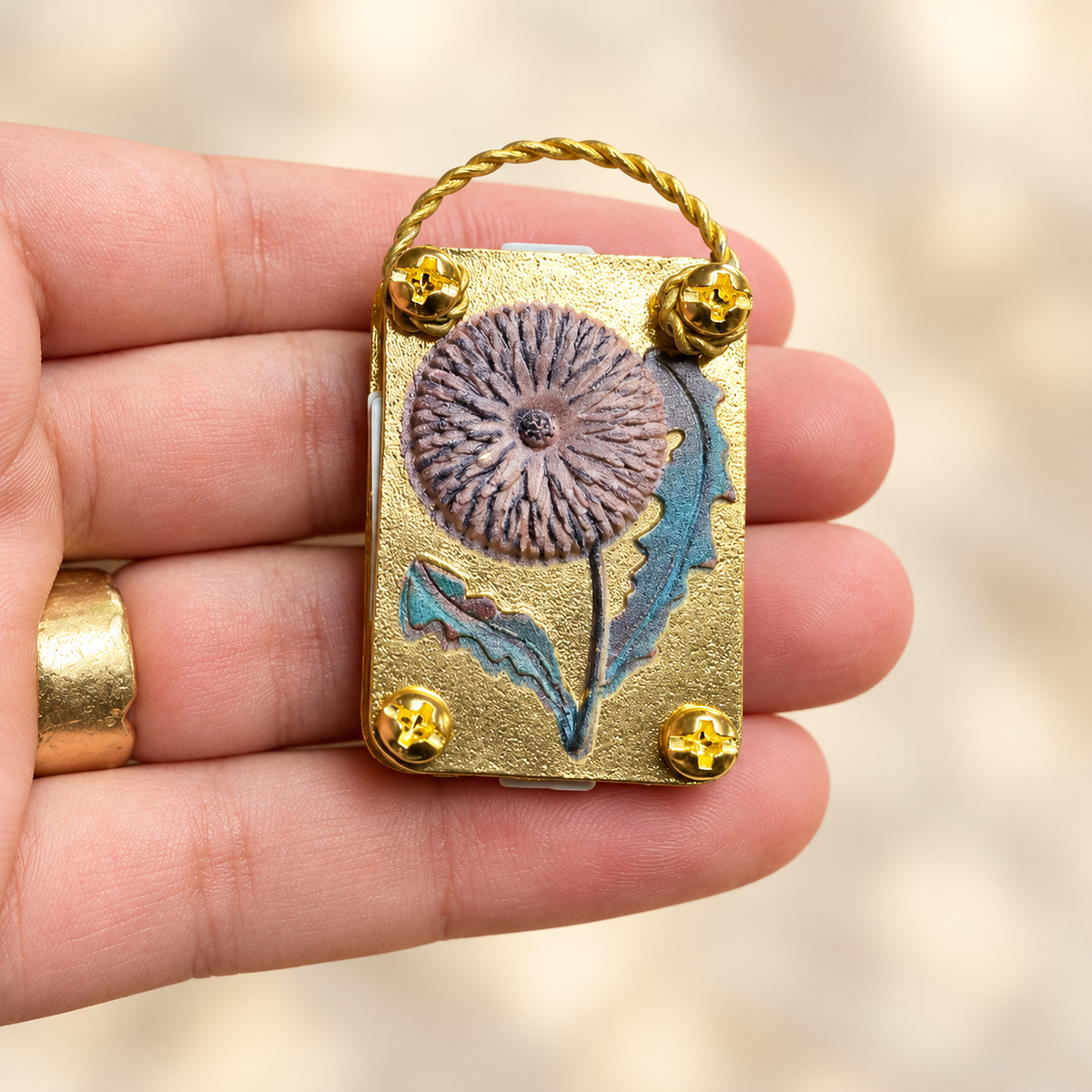 Gold-tone micro flower press with a dandelion design held in a hand for scale; styles vary.